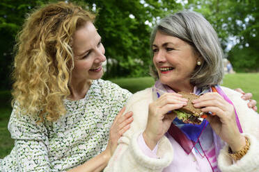 Lächelnde blonde Frau betrachtet ältere Mutter beim Essen eines Sandwiches im Park - ECPF00955