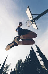 Directly below view of teenage player dunking ball in basketball hoop against sky - GIOF08434