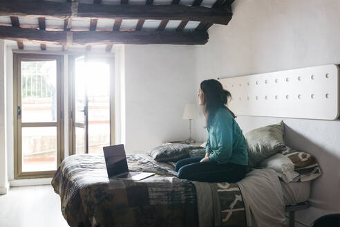 Junge Frau mit Laptop sitzt auf dem Bett und schaut aus dem Fenster - JRFF04513