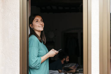 Woman using smartphone at open window, man using laptop lying on bed at home - JRFF04549