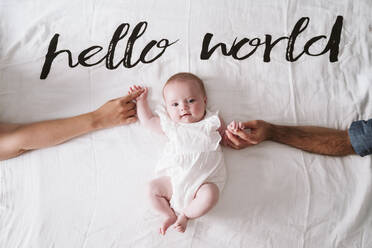 Happy cute baby girl holding hands of parents while lying on blanket under Hello World text at home - EBBF00198