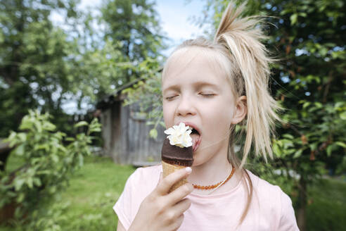 Porträt eines blonden Mädchens mit geschlossenen Augen, das Eis mit Jasminblüten im Garten isst - EYAF01169