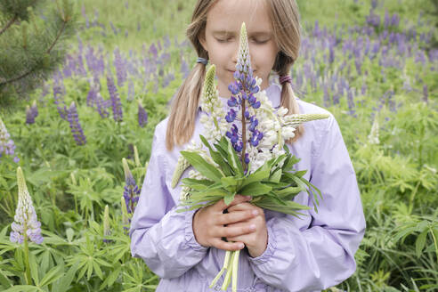 Porträt eines Mädchens mit geschlossenen Augen, das vor einem Blumenfeld steht und einen Strauß Lupinen hält - EYAF01171