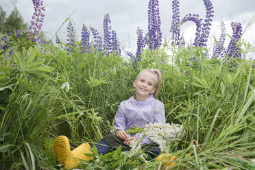 Porträt eines glücklichen Mädchens, das vor einem Lupinenfeld mit einem Strauß gepflückter Blumen sitzt - EYAF01175