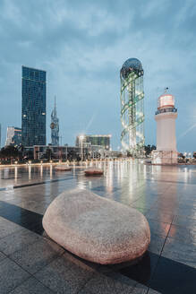 Georgia, Adjara, Batumi, Rock on illuminated town square at dusk with Batumi Lighthouse and Alphabetic Tower in background - WVF01799