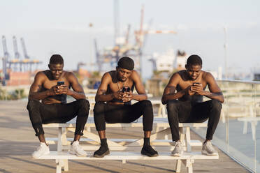 Junge Männer ohne Hemd sitzen nebeneinander auf einem Picknicktisch und benutzen Smartphones an einem sonnigen Tag - EGAF00243
