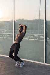 Cheerful shirtless young man enjoying dancing against glass window - EGAF00252