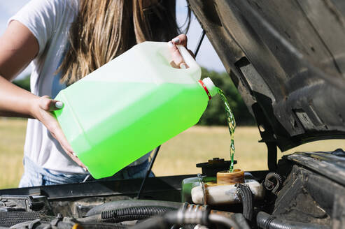 Mittelteil einer Frau auf einer Autofahrt, die an einem sonnigen Tag Kühlmittel in den Motor gießt - JCMF00898