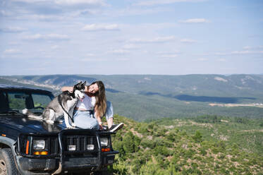 Smiling woman looking at Siberian husky while sitting on 4x4 vehicle hood against sky during road trip - JCMF00900