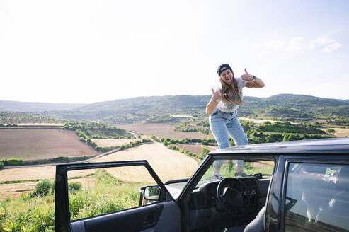 Cheerful woman sticking out tongue while showing hand sign on vehicle hood against sky - JCMF00908