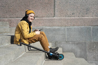 Nachdenklicher Junge hört Musik, während er mit seinem Roller auf einer Treppe sitzt, während eines sonnigen Tages - VPIF02519