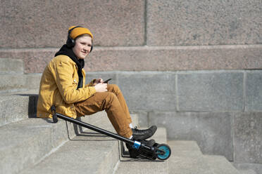 Thoughtful boy listening music while sitting with push scooter on steps during sunny day - VPIF02519