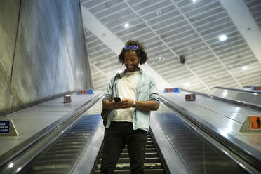 Junger trendiger Mann lächelt, während er sein Smartphone auf einer Rolltreppe in einer U-Bahn-Station benutzt - PMF01113
