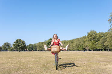 Cheerful woman with feet up riding bicycle on field against clear blue sky - WPEF03025