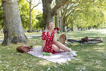 Happy woman holding smart phone while sitting on blanket at public park - WPEF03035