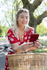 Schöne Frau, die ihr Smartphone benutzt, während sie sich in einem öffentlichen Park auf ein Fahrrad stützt - WPEF03041