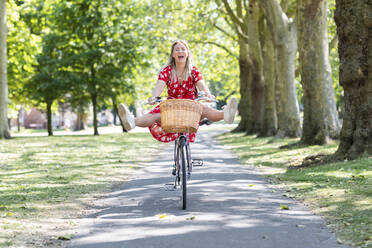 Cheerful woman with feet up riding bicycle on footpath at public park - WPEF03050