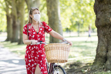 Frau mit Maske, die mit dem Fahrrad auf dem Gehweg in einem öffentlichen Park spazieren geht - WPEF03056