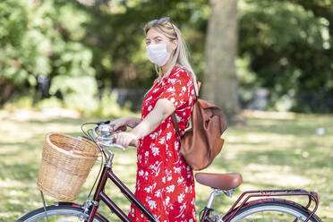 Mittlere erwachsene Frau, die eine Maske trägt, während sie mit dem Fahrrad in einem öffentlichen Park steht - WPEF03058