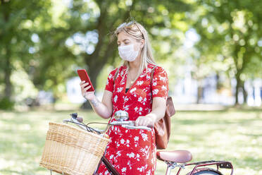 Frau mit Fahrrad trägt Maske, während sie in einem öffentlichen Park ihr Smartphone benutzt - WPEF03061