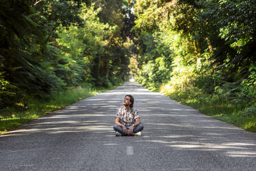 Thoughtful young man sitting on road amidst trees in forest - WVF01821