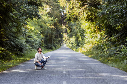 Young man with diary crouching on country road amidst trees stock photo