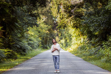 Young man looking up while holding book on country road amidst trees in forest - WVF01829