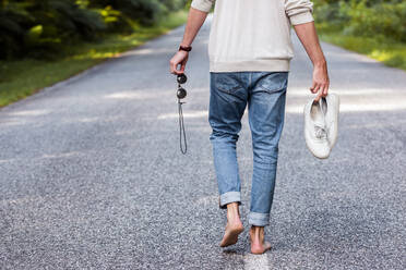 Man holding sports shoes and sunglasses while walking on road in forest - WVF01831