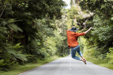 Excited man jumping on country road amidst trees in forest - WVF01833