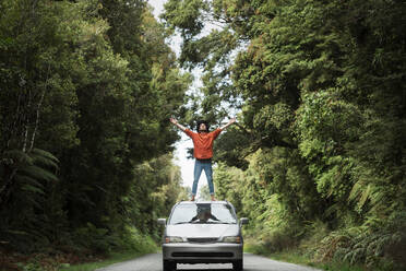 Junger Mann mit ausgestreckten Armen auf dem Autodach stehend an einer Landstraße inmitten von Bäumen - WVF01846