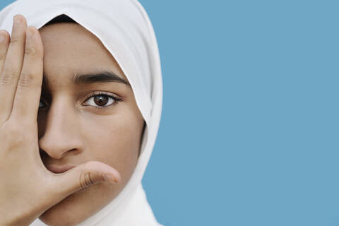 Muslimisches Mädchen bedeckt Auge mit Hand gegen blauen Hintergrund, lizenzfreies Stockfoto