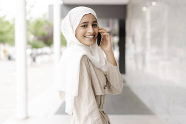 Muslimisches Mädchen im Teenageralter lächelt, während sie über ihr Smartphone in der Stadt spricht - JCZF00157