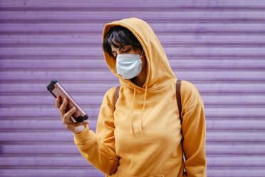 Portrait of young woman with yellow hoodie and protective mask in front of purple background - TCEF00783