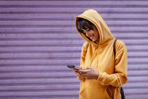 Porträt einer jungen Frau mit gelbem Kapuzenpulli, die ihr Smartphone vor einem lila Hintergrund überprüft - TCEF00787