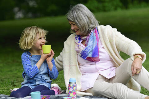 Glückliche Großmutter sieht ihre Enkelin an, die beim Picknick im Park Wasser trinkt, lizenzfreies Stockfoto