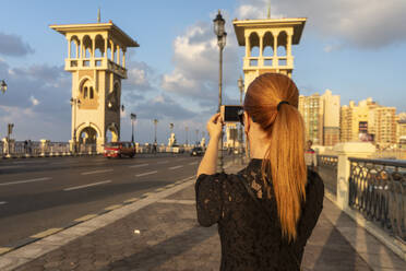 Egypt, Alexandria, Rear view of woman photographing Stanley bridge at sunset - TAMF02330