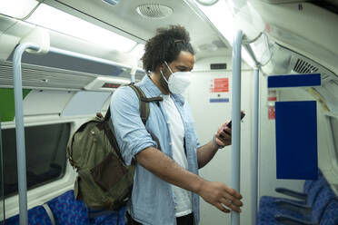 Mann mit Schutzmaske steht im U-Bahn-Zug und schaut auf sein Handy, London, UK - PMF01116