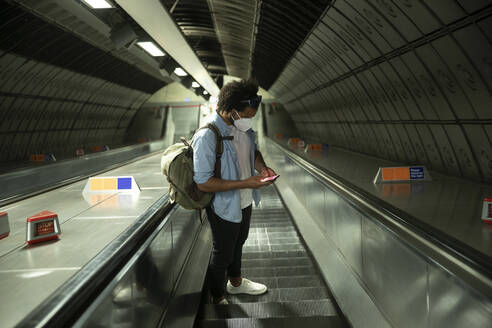Mann mit Schutzmaske steht auf Rolltreppe und schaut auf sein Handy, London, UK - PMF01117