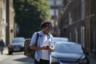Lächelnder junger Mann mit Rucksack, der in einer Wohnstraße steht und auf sein Handy schaut, London, Großbritannien - PMF01119