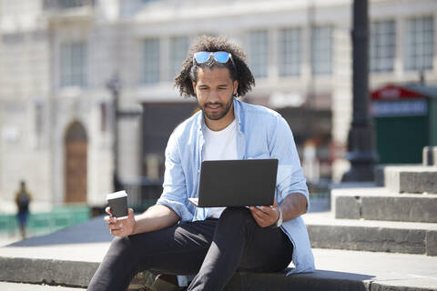 Porträt eines jungen Mannes mit Kaffee zum Mitnehmen, der auf einer Treppe im Freien sitzt und auf einen Laptop schaut, London, UK, lizenzfreies Stockfoto