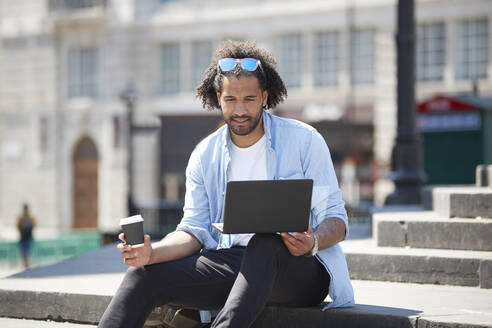 Porträt eines jungen Mannes mit Kaffee zum Mitnehmen, der auf einer Treppe im Freien sitzt und auf einen Laptop schaut, London, UK - PMF01128