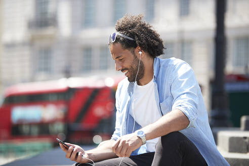 Porträt eines lächelnden jungen Mannes, der im Freien sitzt und mit Handy und Kopfhörern Musik hört, London, UK - PMF01130