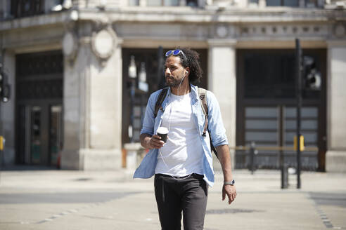 Porträt eines jungen Mannes mit Kaffee zum Mitnehmen, der die Straße überquert, während er mit Kopfhörern Musik hört, London, UK - PMF01132