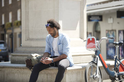 Junger Mann mit Leihfahrrad hört Musik mit Handy und Kopfhörern in der Stadt, London, UK, lizenzfreies Stockfoto