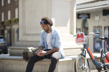 Junger Mann mit Leihfahrrad hört Musik mit Handy und Kopfhörern in der Stadt, London, UK - PMF01139