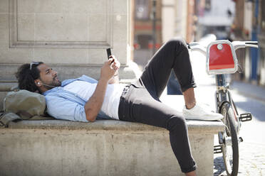 Junger Mann mit Leihfahrrad, der mit Kopfhörern Musik hört und auf sein Handy schaut, London, UK - PMF01140