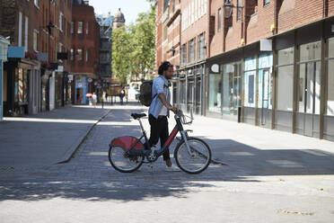 Junger Mann beim Überqueren der Straße mit Leihfahrrad, London, UK - PMF01142