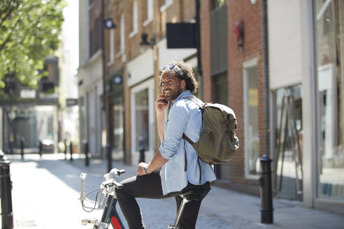 Porträt eines lächelnden jungen Mannes am Telefon mit Leihfahrrad und Rucksack in der Stadt, London, UK - PMF01143