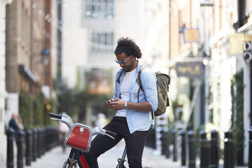 Junger Mann mit Leihfahrrad und Rucksack, der sein Handy in der Stadt benutzt, London, Großbritannien - PMF01144