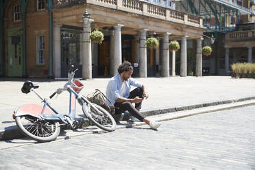 Junger Mann mit Leihfahrrad und Rucksack sitzt am Straßenrand und schaut auf sein Handy, London, Großbritannien - PMF01147